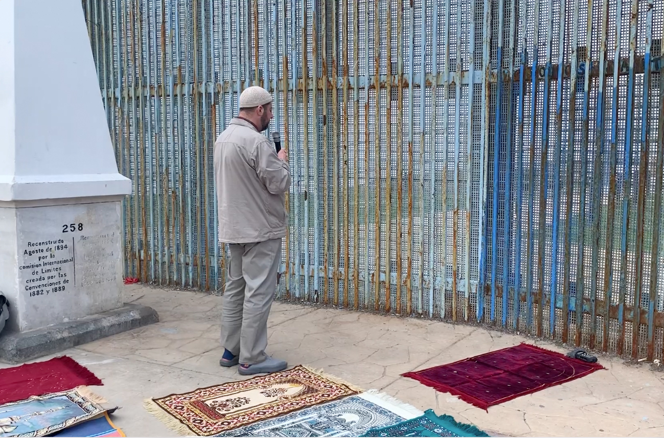 A man in a beige jacket, pants, and skull cap speaks into a microphone while facing a tall blue metal fence. Rectangular prayer rugs lie on the ground below him
